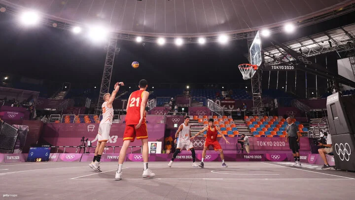 A basketball player in a gray jersey is jumping and attempting a layup, holding a blue and yellow basketball close to the hoop. Another player in front is defending, with one arm raised. A third player is behind, also attempting to block the shot. The background shows an audience seated in a stadium, with some people wearing masks.