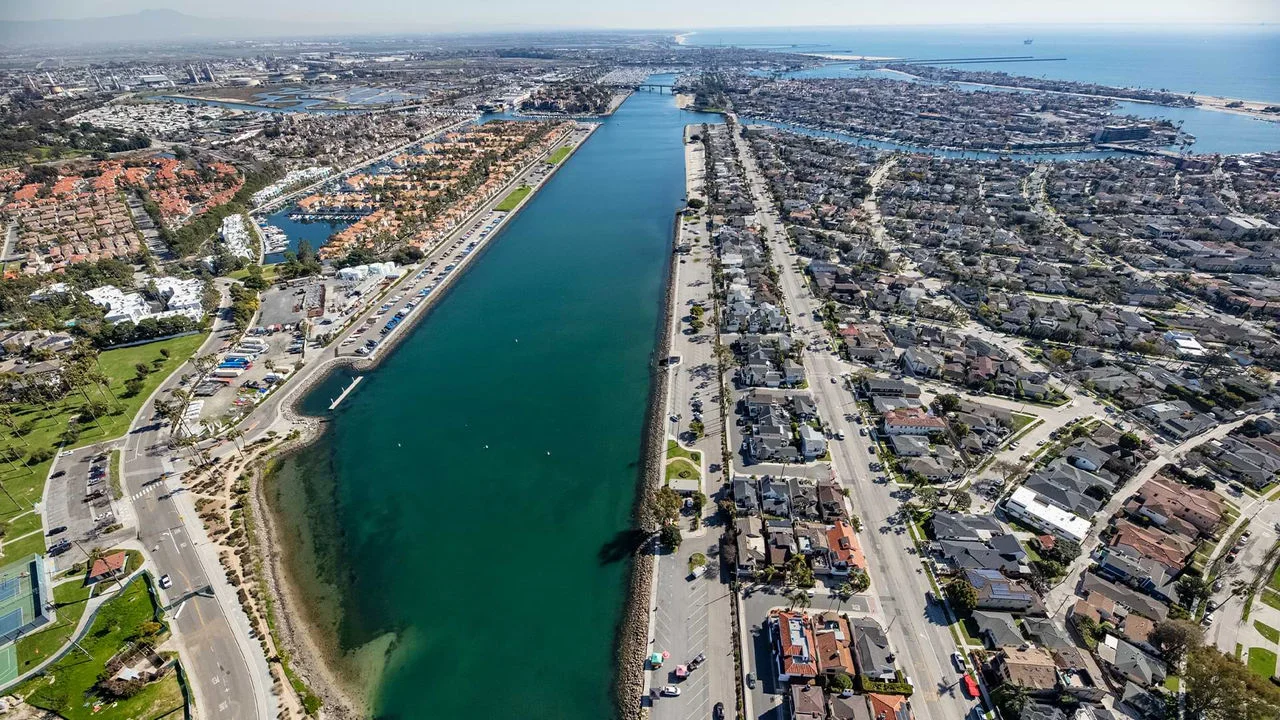 Aerial photograph showing a large rectangular body of blue and green water with roads, building and houses bordering both sides. The body of water stretches back to an outlet that connects to the ocean which can be seen in the background below a blue sky.