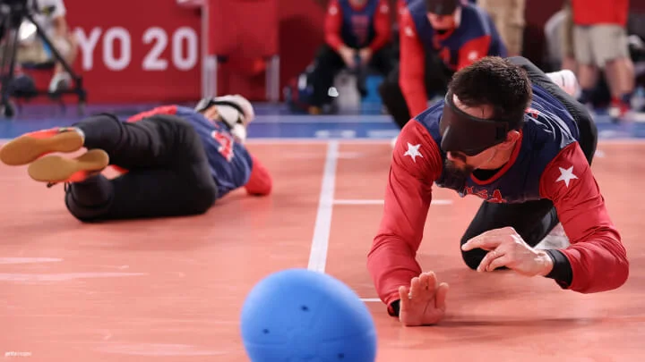 Dos jugadores de goalball están en la cancha, ambos usando antifaces. El jugador en primer plano, vestido con una camiseta azul y roja con estrellas blancas en los hombros, está en una posición defensiva baja con las manos extendidas hacia un goalball azul. El jugador en el fondo, vestido igualmente con una camiseta azul y roja, descansa de lado en el suelo. La escena se desarrolla en una arena cubierta con un piso rojo y un letrero parcialmente visible de "Tokio 2020" en el fondo.