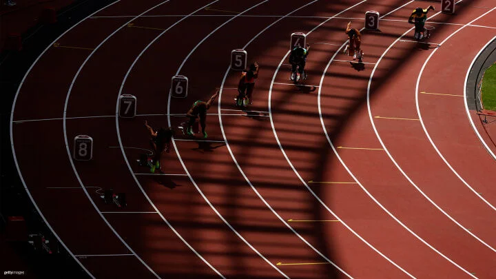 Se capta a un grupo de velocistas en los bloques de partida de una carrera en una pista curvada bajo una iluminación dramática. Las sombras de la estructura del estadio crean un patrón sobre la pista. Cada corredor está en el bloque de salida, lanzándose hacia adelante. Los números de los carriles del 2 al 8 están claramente visibles.