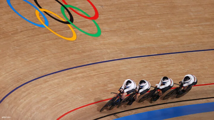 Un grupo de cuatro ciclistas, vestidos con uniformes blancos idénticos y cascos aerodinámicos, pedalean juntos en fila sobre una pista del velódromo de madera cubierta. Están recorriendo una curva muy inclinada, marcada con líneas rojas, azules y negras. Sobre ellos, los anillos del logo Olímpico son visibles en la superficie de madera.