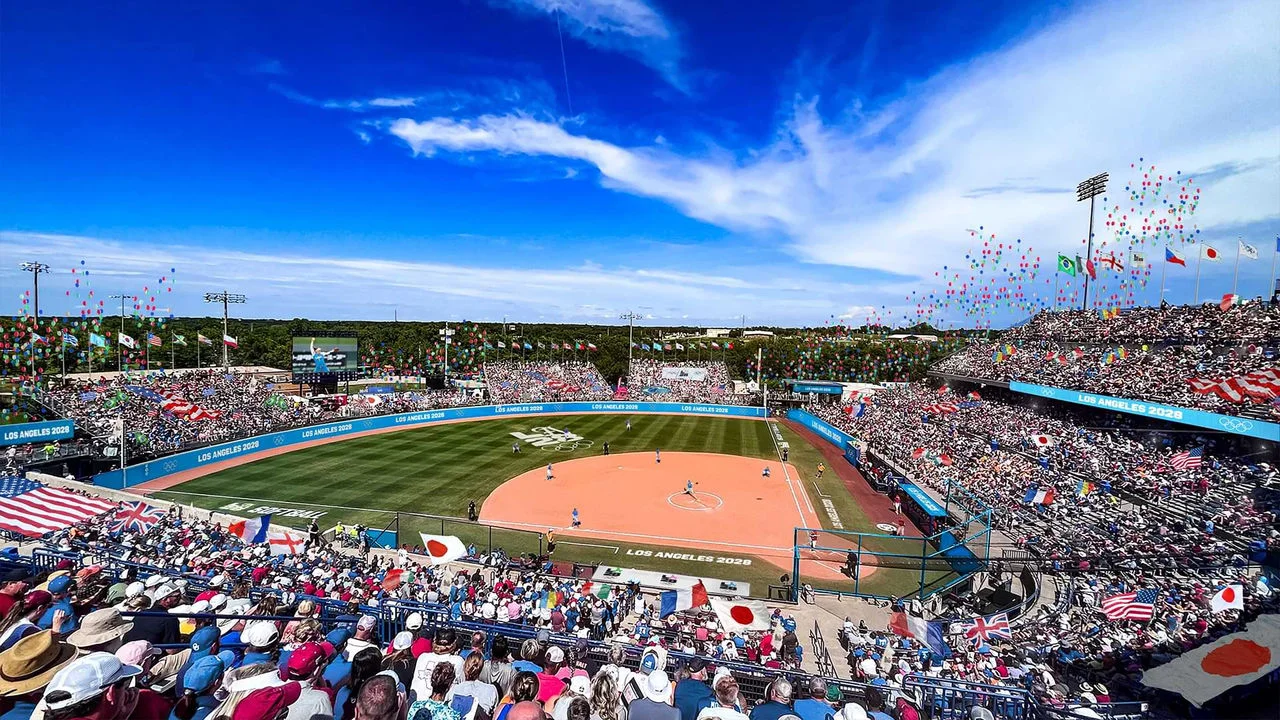 A digital rendering shows a softball game at the Olympic Softball Park, also known as Devon Park. The field and screens display "Los Angeles 2028" and the LA28 logo. The stadium is full of cheering fans, many waving flags from different countries. Colorful balloons float in the sky above. The green field is well-maintained with players actively participating in the game.