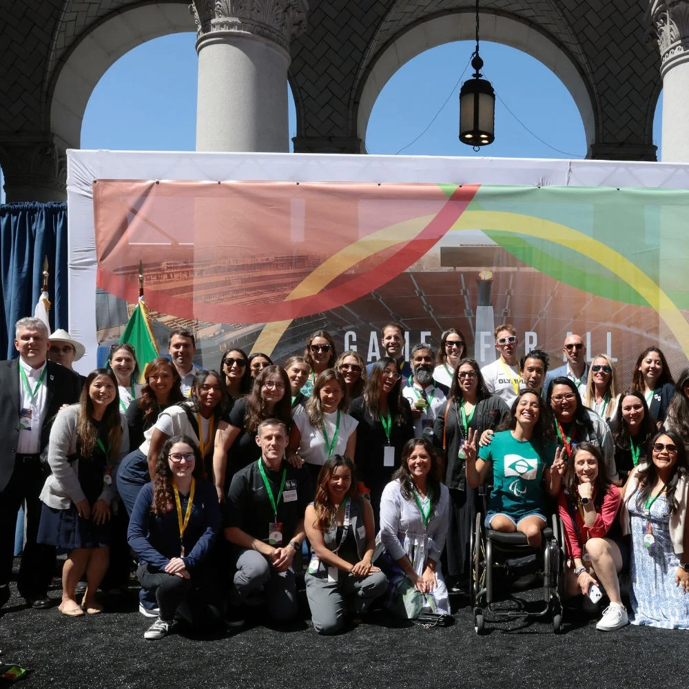 Fotografía mostrando miembros del personal de la organización LA28 en el escenario durante la ceremonia de instalación de la bandera olímpica y paralímpica en el Ayuntamiento de Los Ángeles.