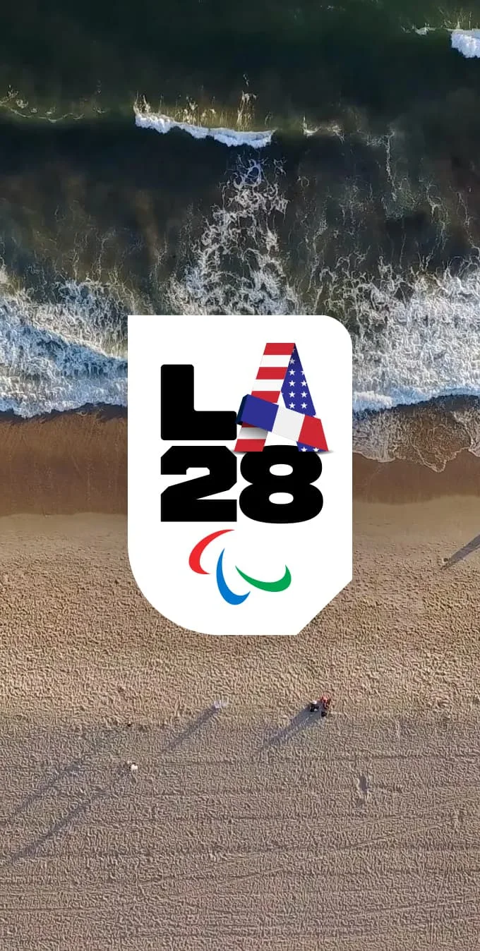 Aerial photograph of a Los Angeles beach with sand and the waves and the LA28 Paralympic Handover A Emblem placed on top of the image on a white background. 