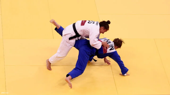 Two judo athletes are competing on a yellow mat. One athlete, wearing a white judogi and black belt, is performing a throw on their opponent, who is wearing a blue judogi and black belt. The athlete in white is leaning over the athlete in blue, gripping their uniform, while the athlete in blue is being lifted off the mat, their right hand touching the ground for support. Both athletes have identifying text on the back of their uniforms.