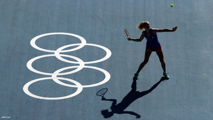 Una jugadora femenina de tenis es capturada en plena acción en una cancha azul, preparándose para golpear una pelota de tenis en el aire. Los anillos olímpicos se muestran de manera prominente en la cancha a su izquierda. La iluminación proyecta una sombra clara de la jugadora en la cancha, resaltando su postura y la raqueta elevada. Viste una camiseta sin mangas azul, pantalones cortos rojos y zapatos turquesa.