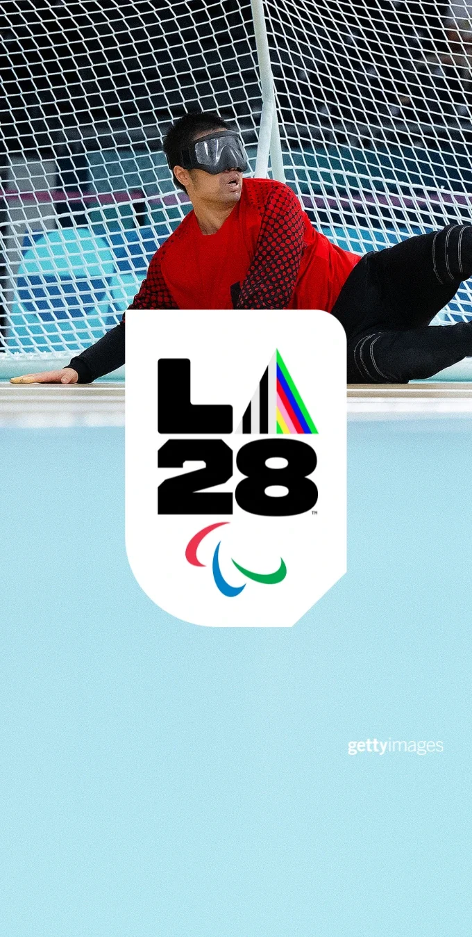 Una fotografía muestra a jugadores paralímpicos de goalball alcanzando un balón azul con una portería de fondo. El emblema del Prisma 'A' de LA28 está superpuesto sobre la imagen. 