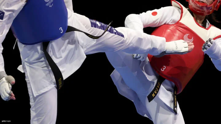 A close-up of two Paralympic taekwondo athletes captured against a black background mid-fight during a competition. The athlete on the left is wearing a blue chest guard and a white uniform with a black belt, delivering a high kick. The athlete on the right, wearing a red chest guard and a white uniform with Japan’s flag and a black belt, is blocking the kick. Both athletes are wearing protective headgear and gloves.