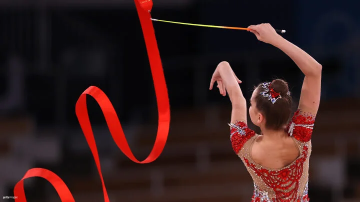 A rhythmic gymnast performs with a red ribbon against a blurred background. She is dressed in a sparkly red and nude leotard adorned with intricate designs. Her hair is neatly tied up in a bun, decorated with a matching red and silver accessory. She holds the ribbon stick high, creating loops and curves with the flowing ribbon.