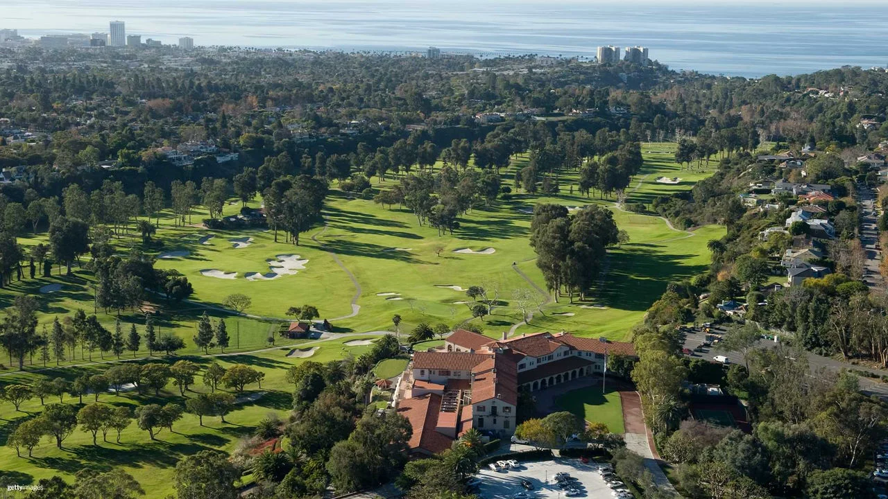 An aerial photograph of the Riviera Country Club showing a large tan building with a red brick roof and behind it an expansive golf course with bright green grass, numerous green trees and foliage surrounding the course, as well as buildings and homes bordering the golf course with ocean views in the background and tall buildings.