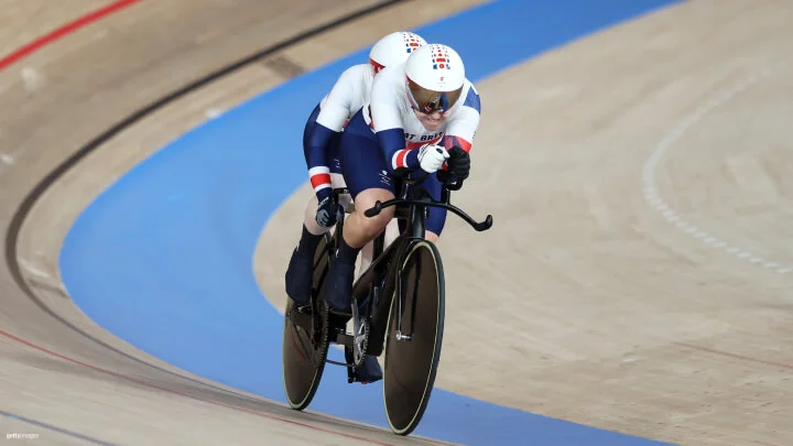 Two cyclists are riding a tandem bike on an indoor velodrome track. Both athletes are dressed in matching white, blue, and red cycling suits from Team Great Britain, with white aerodynamic helmets and goggles. The front cyclist is gripping the handlebars tightly, while the cyclist at the back is slightly obscured. They are leaning forward in an aerodynamic position as they speed around the curved section of the track, which features a light wooden surface with blue and red markings. The background shows the smooth, banked curves of the velodrome.