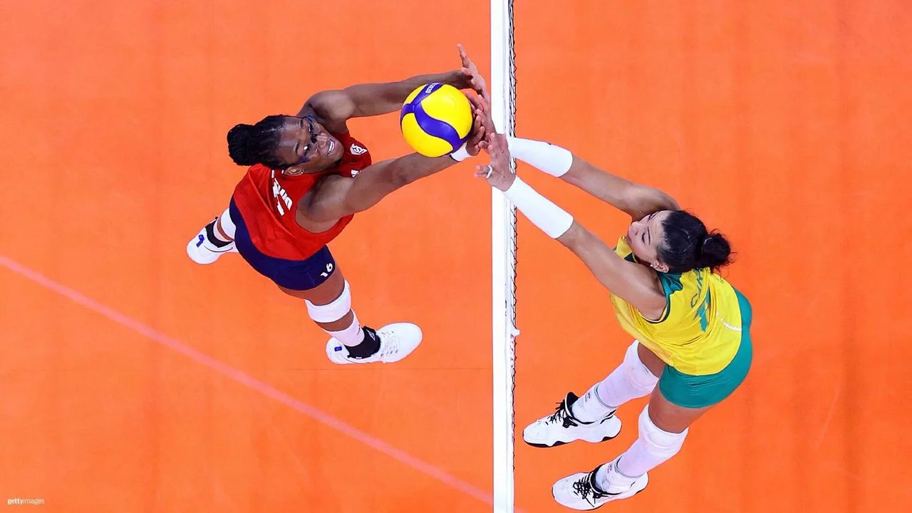 An overhead shot of two volleyball players mid-air at the net, competing for the ball. The player on the left, wearing a red jersey and blue shorts from Team USA, extends her arms to block or spike the ball. The player on the right, from the Brazil team, wearing a yellow and green uniform, also reaches up to block or spike. The volleyball is yellow and blue. The court beneath them is bright orange.