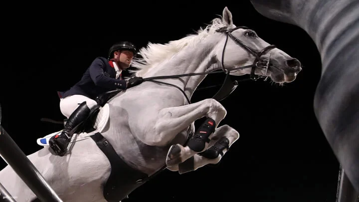 Un jinete masculino, vestido en una chaqueta azul marino, pantalones blancos y un casco negro dirige a un caballo blanco sobre un obstáculo alto en un evento nocturno. El caballo está saltando con las patas delanteras recogidas y la melena ondeando. El jinete se inclina hacia adelante, sujetando las riendas.