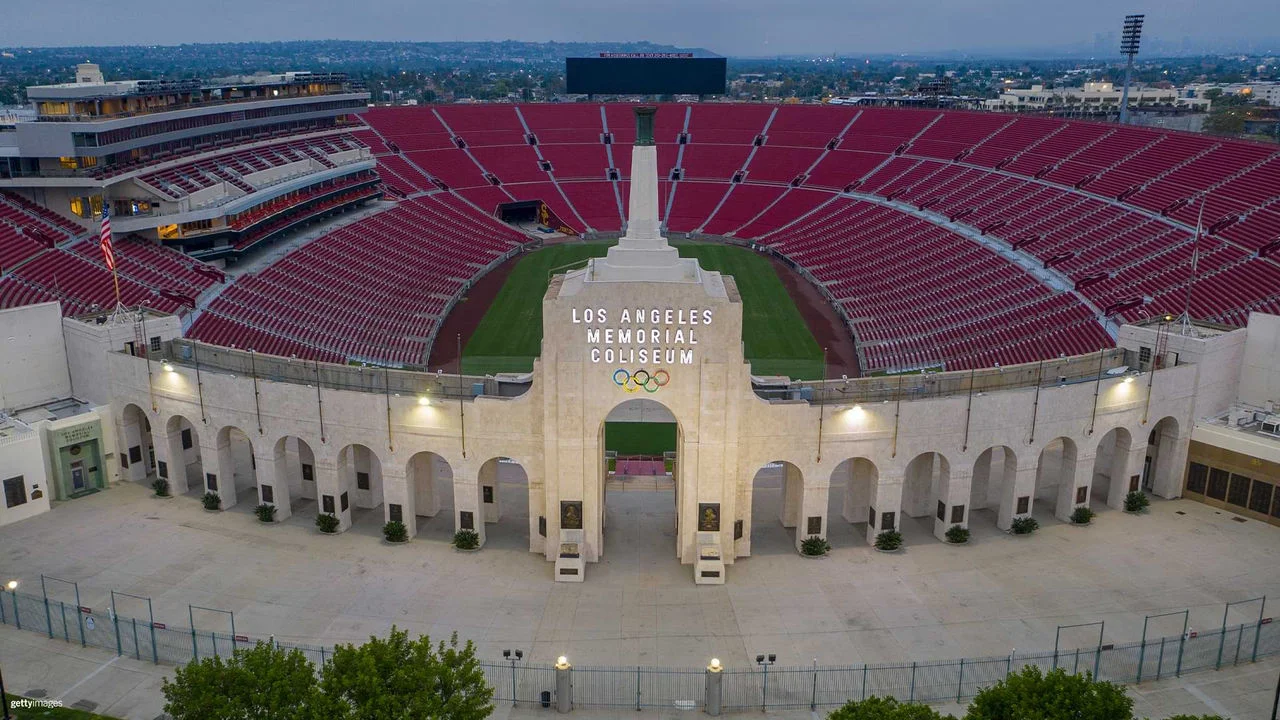 Fotografía del LA Memorial Coliseum desde el frente al atardecer, con los asientos rojos visibles en el fondo, parte del campo verde y las puertas delanteras de color beige con luces encendidas sobre los arcos de piedra.