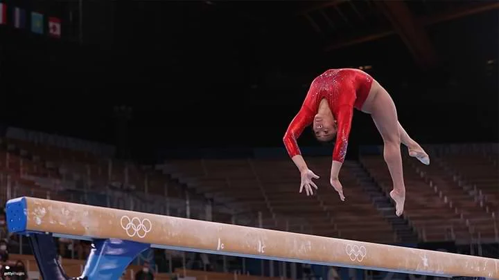 Fotografía de una gimnasta femenina en un maillot rojo haciendo un salto hacia atrás en una barra, capturada en pleno aire.