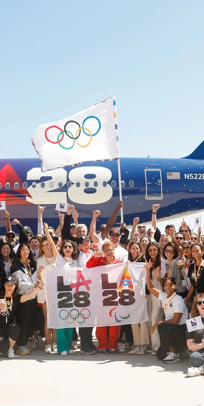 Fotografía mostrando el personal de LA28 sosteniendo la bandera de los anillos olímpicos y la bandera de LA28 en frente del avión de Delta del vuelo de la bandera olímpica