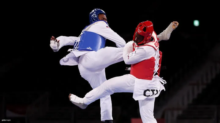 Dos atletas con equipo de protección están en un combate de taekwondo. El atleta a la izquierda, con equipo de protección azul, está realizando una patada alta hacia el atleta a la derecha, que lleva equipo de protección rojo. Ambos atletas están en el aire. 