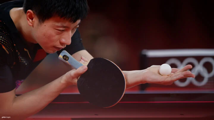 Una imagen cercana de un jugador masculino de tenis de mesa con una camiseta negra, intensamente concentrado mientras se prepara para servir. Sostiene una raqueta en su mano derecha y una pelota en su mano izquierda, que está extendida hacia afuera. El fondo está borroso, pero se pueden ver los anillos olímpicos en la red.