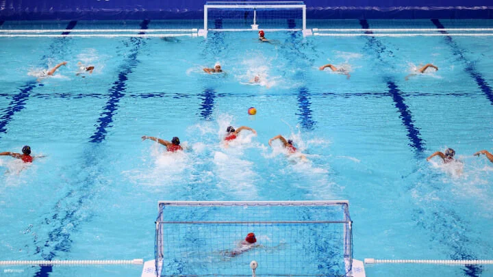 A water polo match is in progress in a large, bright blue pool. Two teams are seen swimming vigorously towards the ball, which is floating in the center of the pool. Players are wearing different colored caps to distinguish the teams, with the goalkeepers positioned at either end of the pool, each guarding a goal. The action is intense, with players creating splashes as they swim and prepare to compete for control of the ball.