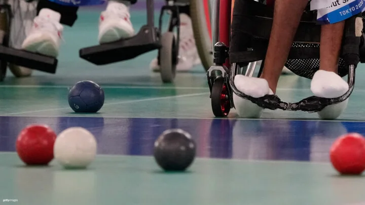 Una imagen cercana de un juego de boccia, enfocándose en la parte inferior de dos atletas en sillas de ruedas. El deportista en la silla de ruedas de color rojo tiene los pies apoyados en los reposapiés y lleva calcetines blancos. El atleta a la izquierda es parcialmente visible en el fondo borroso, con zapatillas blancas descansando sobre el reposapiés. Varias bolas de boccia, incluyendo rojas, azules, blancas y negras, están dispersas en la cancha.