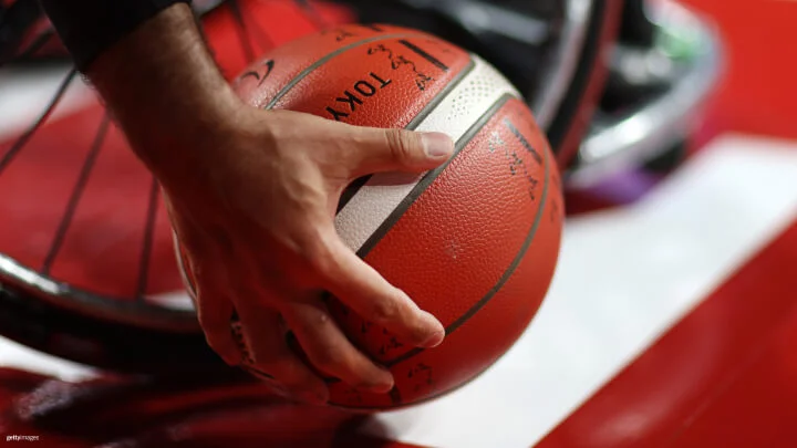 Una imagen cercana de una mano agarrando un balón de baloncesto. El balón es de color naranja con líneas negras y blancas y tiene la palabra "Tokio" impresa en él. En el fondo se ve la rueda de una silla de ruedas, indicando que el contexto está vinculado al baloncesto en silla de ruedas. La atención se centra en la mano que sujeta el balón de baloncesto, mientras la silla de ruedas aparece ligeramente desenfocada al fondo