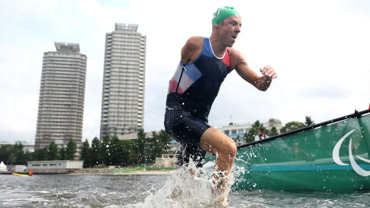 A Paralympic athlete, wearing a triathlon suit and a green swim cap, is emerging from the water with droplets splashing around him. The athlete has his right arm amputated at the shoulder. In the background, the sky is overcast, and there are two tall buildings and some greenery.