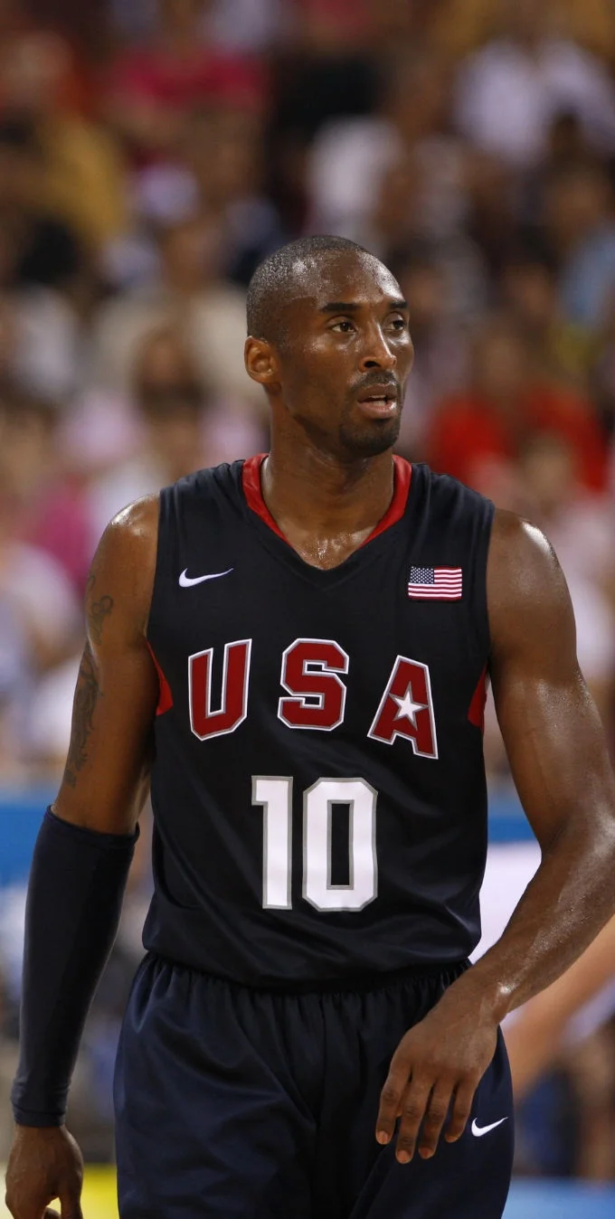 A photograph of Kobe Bryant wearing a navy Team USA basketball jersey while playing basketball in a stadium with a crowd in the background. 