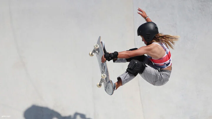 Una skater femenina es capturada en el aire mientras realiza un truco en un skatepark. Ella lleva puesto un casco negro, una camiseta sin mangas colorida, pantalones grises y protecciones en las muñecas y las rodillas. Con el cabello largo fluyendo hacia atrás, sostiene su tabla de skate con las rodillas flexionadas y una mano extendida para mantener el equilibrio. El fondo es una superficie de concreto liso y claro.