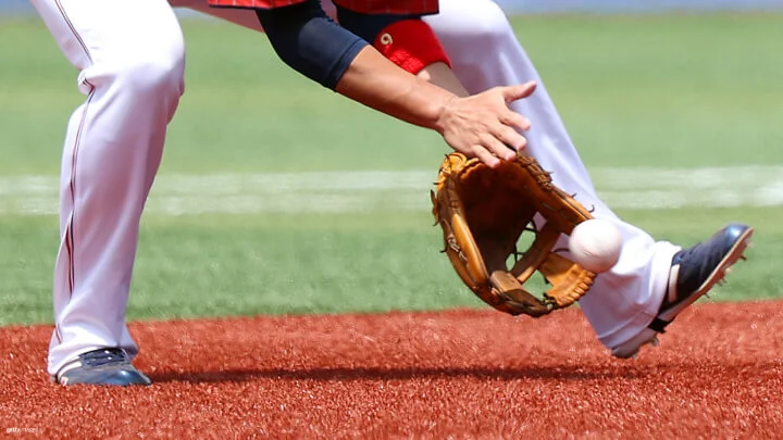 A baseball player wearing white pants, a red jersey, and black cleats is in the process of fielding a ground ball. The player's gloved hand is positioned low to the ground, just about to catch the baseball, while the player's other hand is placed nearby, ready to secure the ball. The scene takes place on a baseball field with a green turf background and a reddish-brown infield.